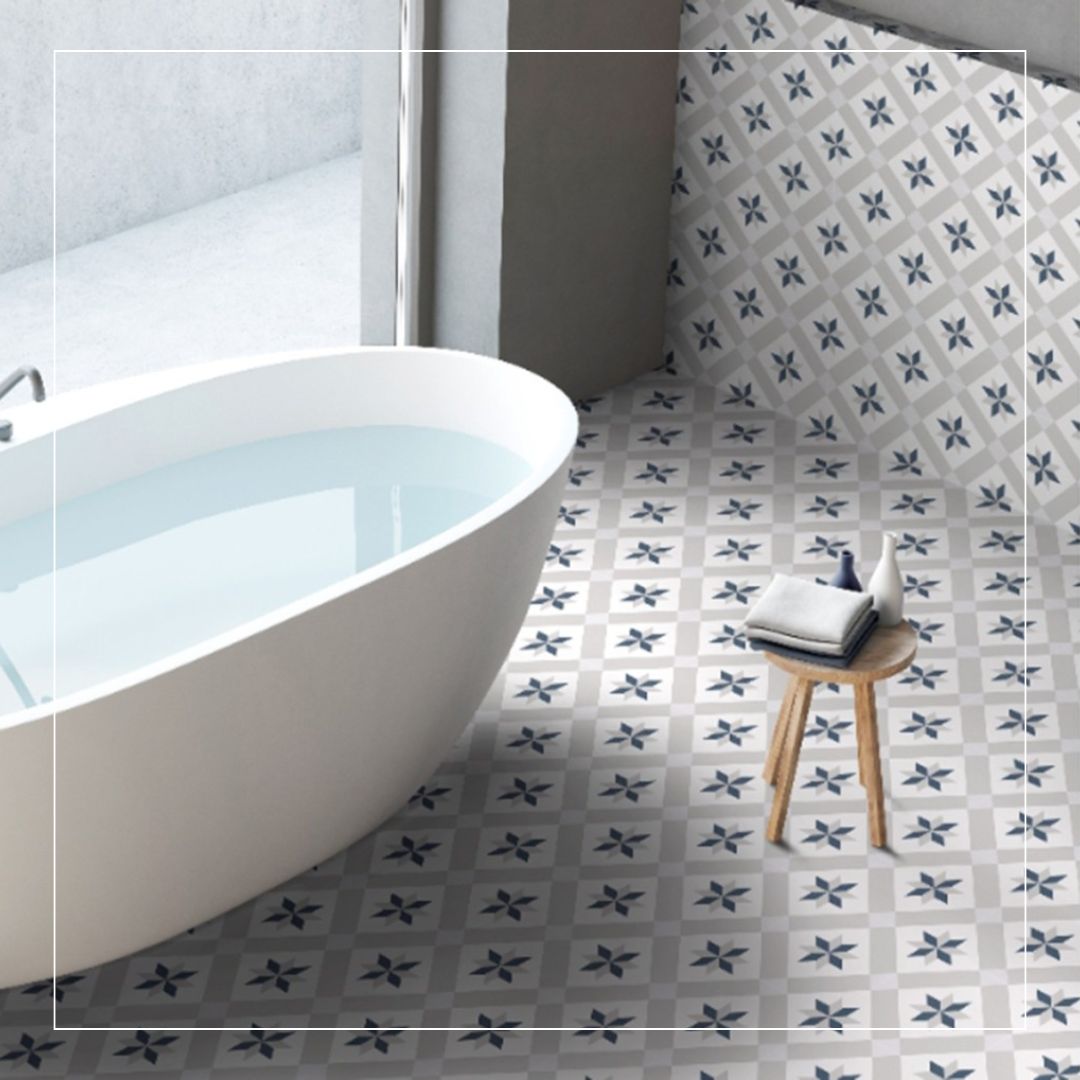 Modern bathroom with a freestanding white bathtub and blue and grey patterned floor tiles featuring a geometric floral motif, styled with a wooden stool and folded towels.