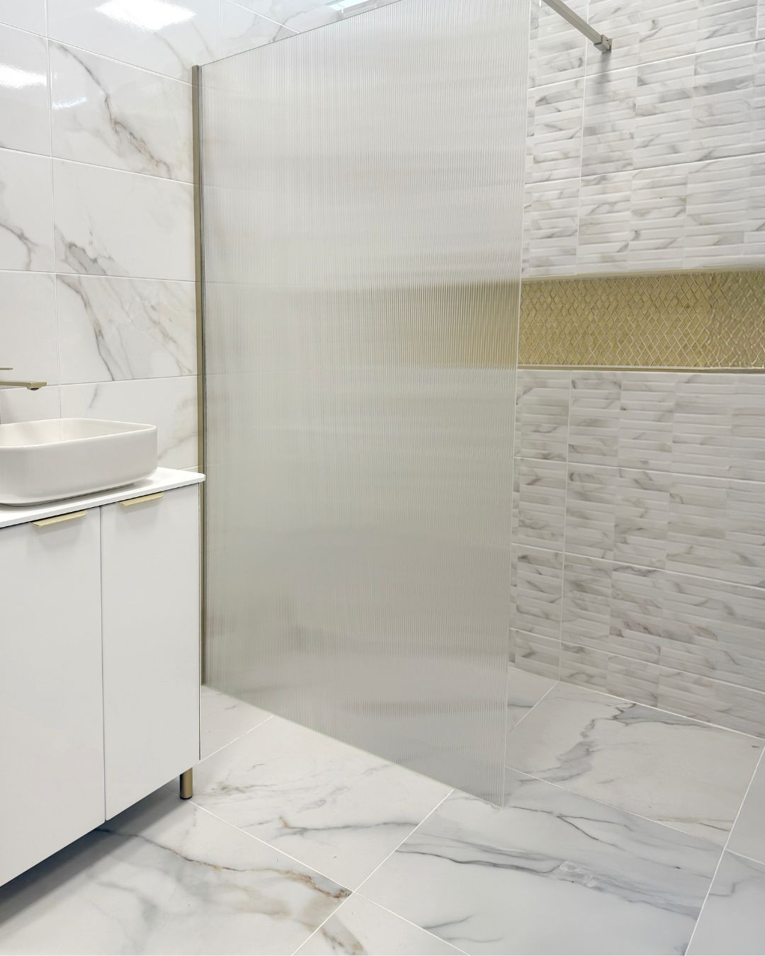 Contemporary wet room with large white marble-effect floor tiles, matching wall tiles, fluted glass shower screen and gold mosaic feature tiles