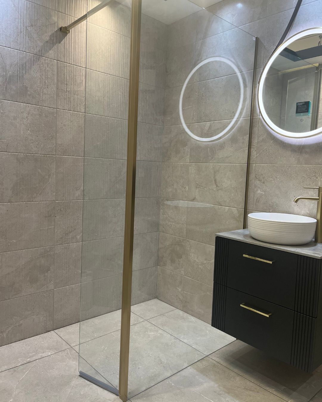 Modern wet room with large grey stone-effect floor and wall tiles, walk-in shower with glass screen, black floating vanity unit and brushed gold fittings.
